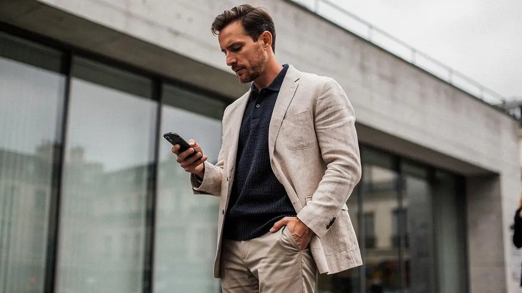 Homme portant un polo en maille à manches longues sous un blazer non structuré, consultant son téléphone devant un bâtiment contemporain